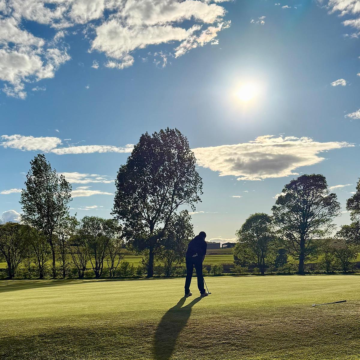 A golfer putting on a green, silhouetted against a bright sun with scattered clouds and trees in the background.