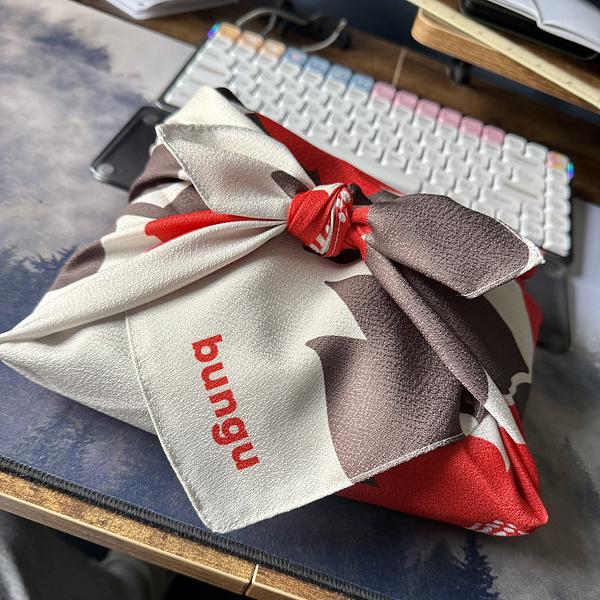 A furoshiki-style cloth wrapping in white, red, and brown with the text "bungu" printed in red, tied in a knot on a desk next to a white mechanical keyboard and smartphone.