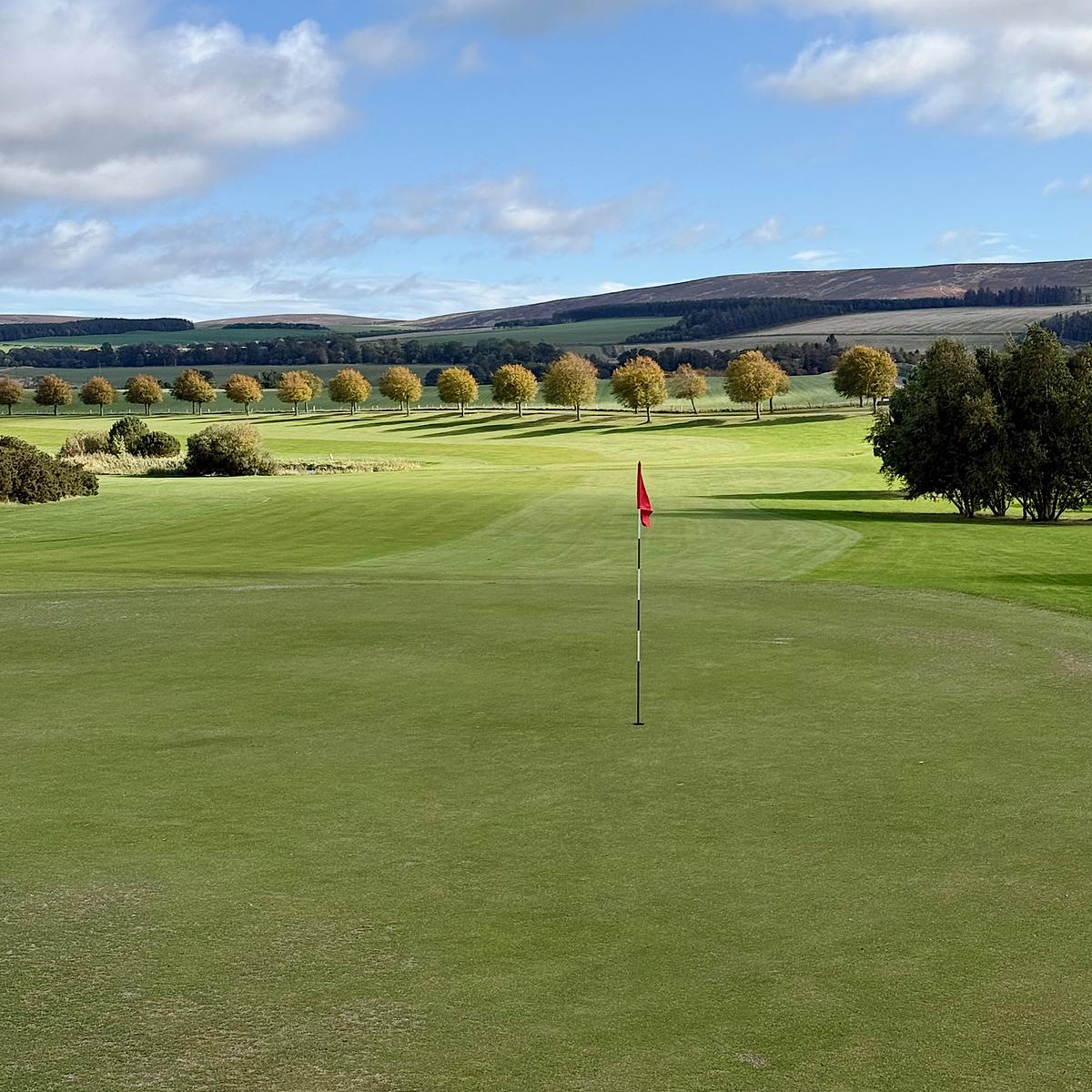 Golf course green with red flag, lined by autumn-colored trees and rolling green hills under a partly cloudy sky.