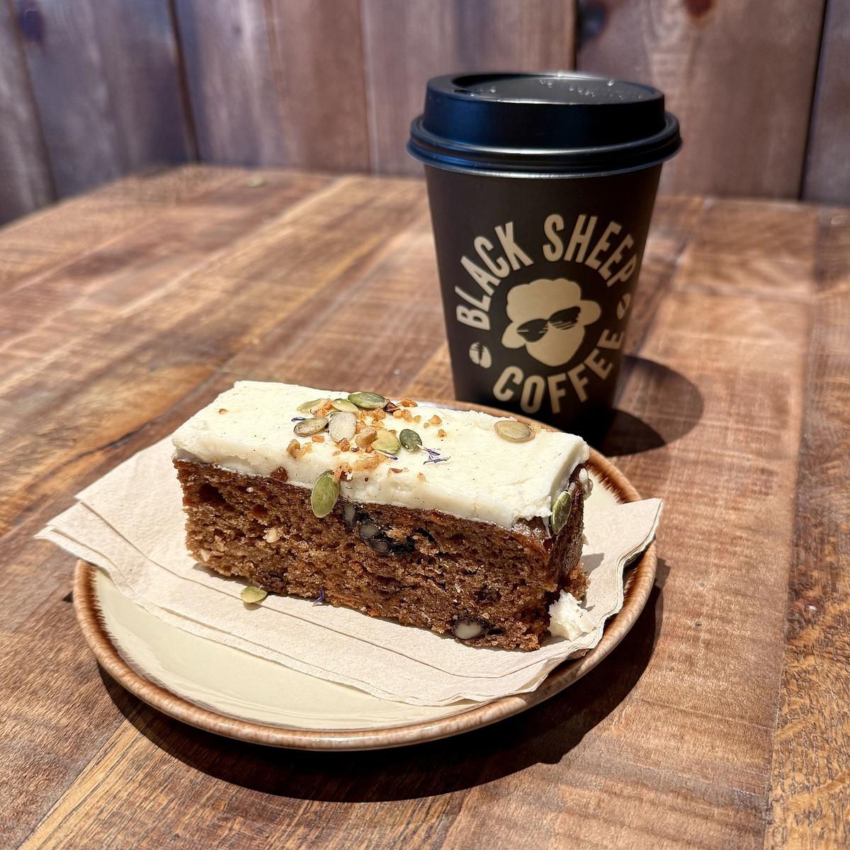 A slice of cake with frosting and seeds is served on a plate next to a takeaway cup of Black Sheep Coffee.