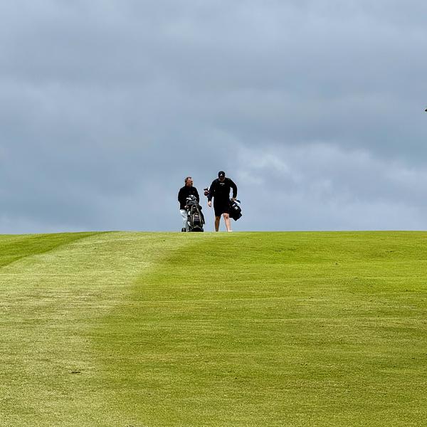 Two golfers walking on a green hilltop of a golf course under overcast skies, carrying their golf bags.