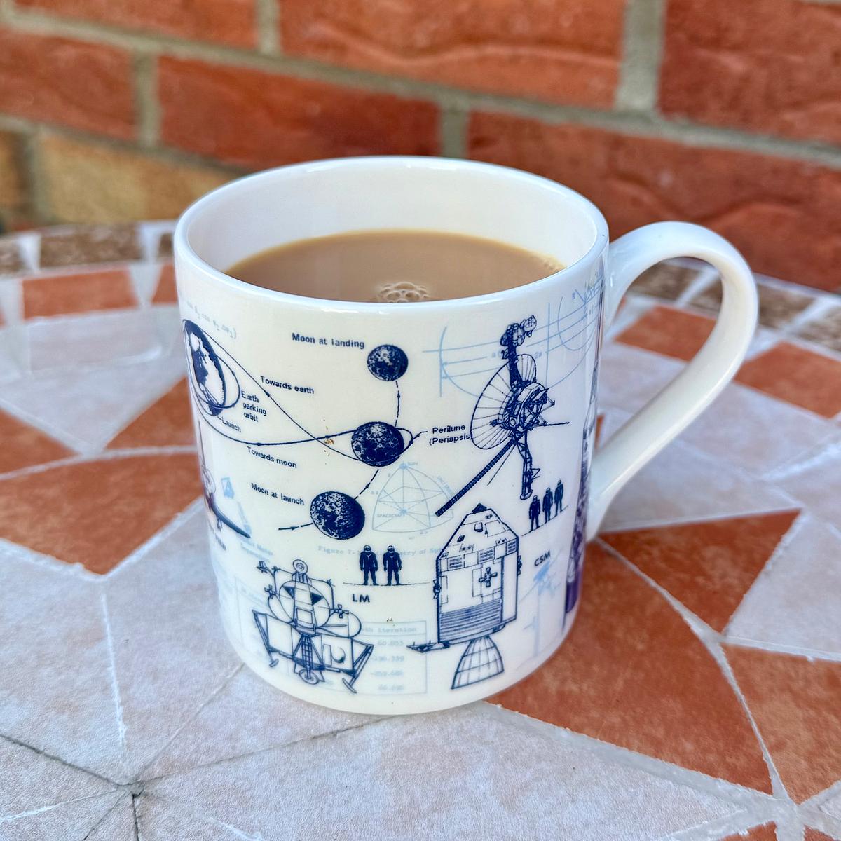 White ceramic mug filled with tea, decorated with blue space exploration diagrams including lunar orbit trajectories and spacecraft blueprints.