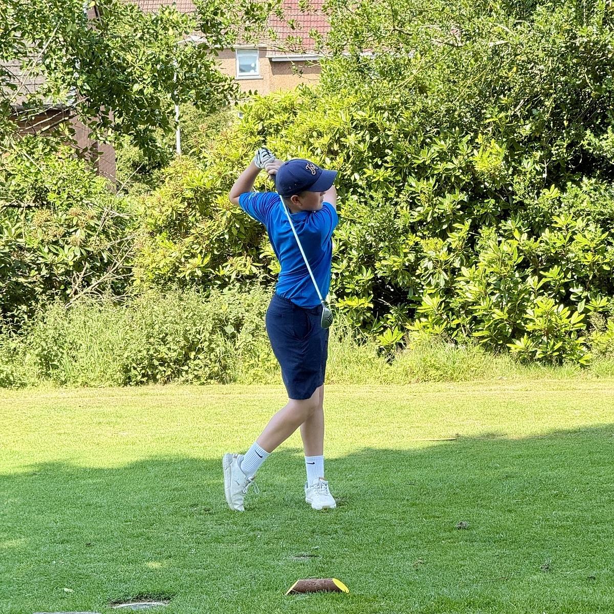 A person is playing golf, swinging a club on a grassy course with trees in the background.