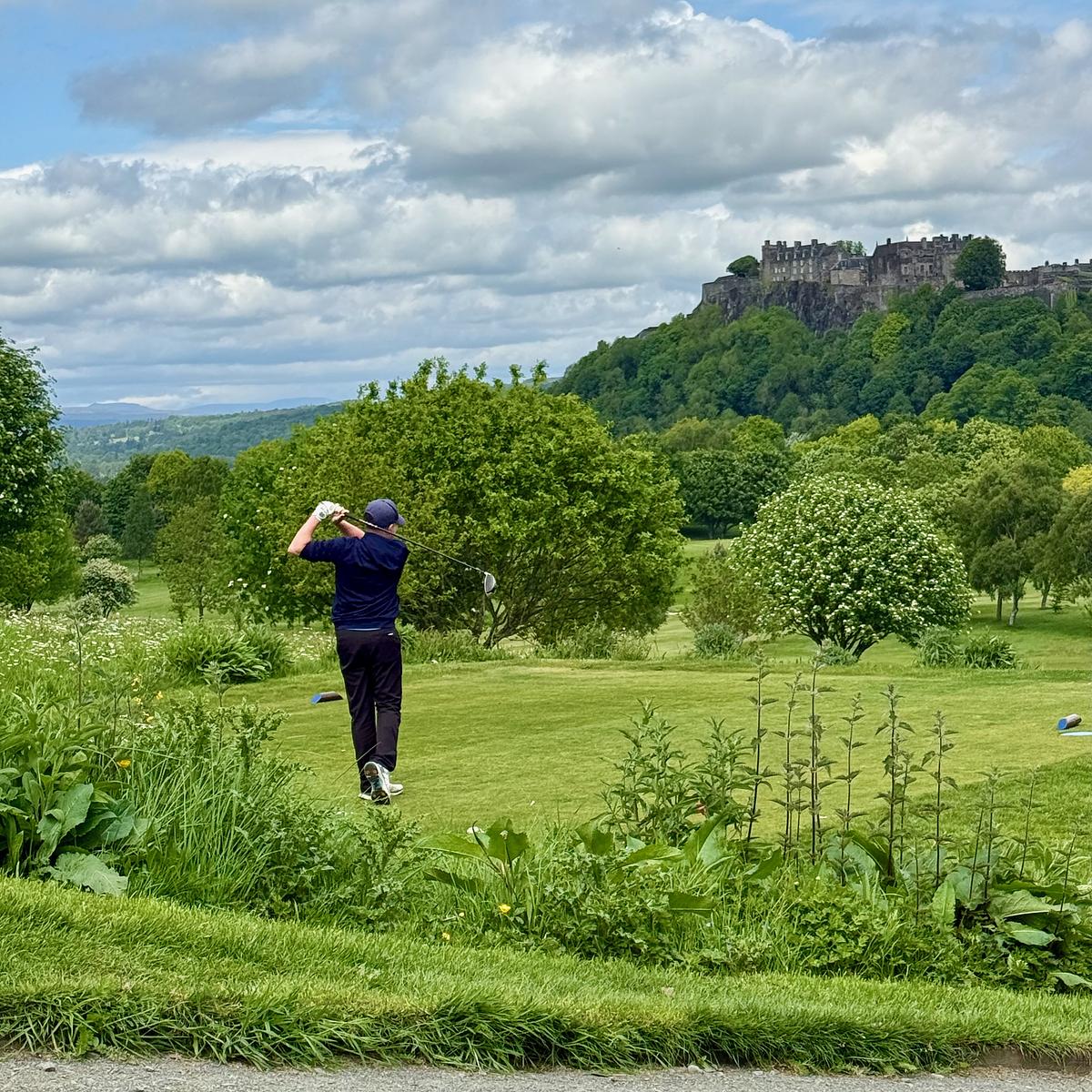 A golfer mid-swing on a lush green course with Stirling Castle perched on a hilltop in the background under cloudy skies.