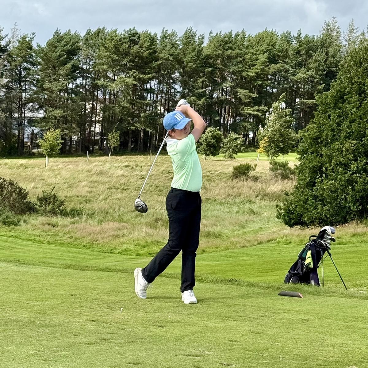 A golfer in a green shirt swings a club on a lush golf course, with trees and a clear sky in the background.