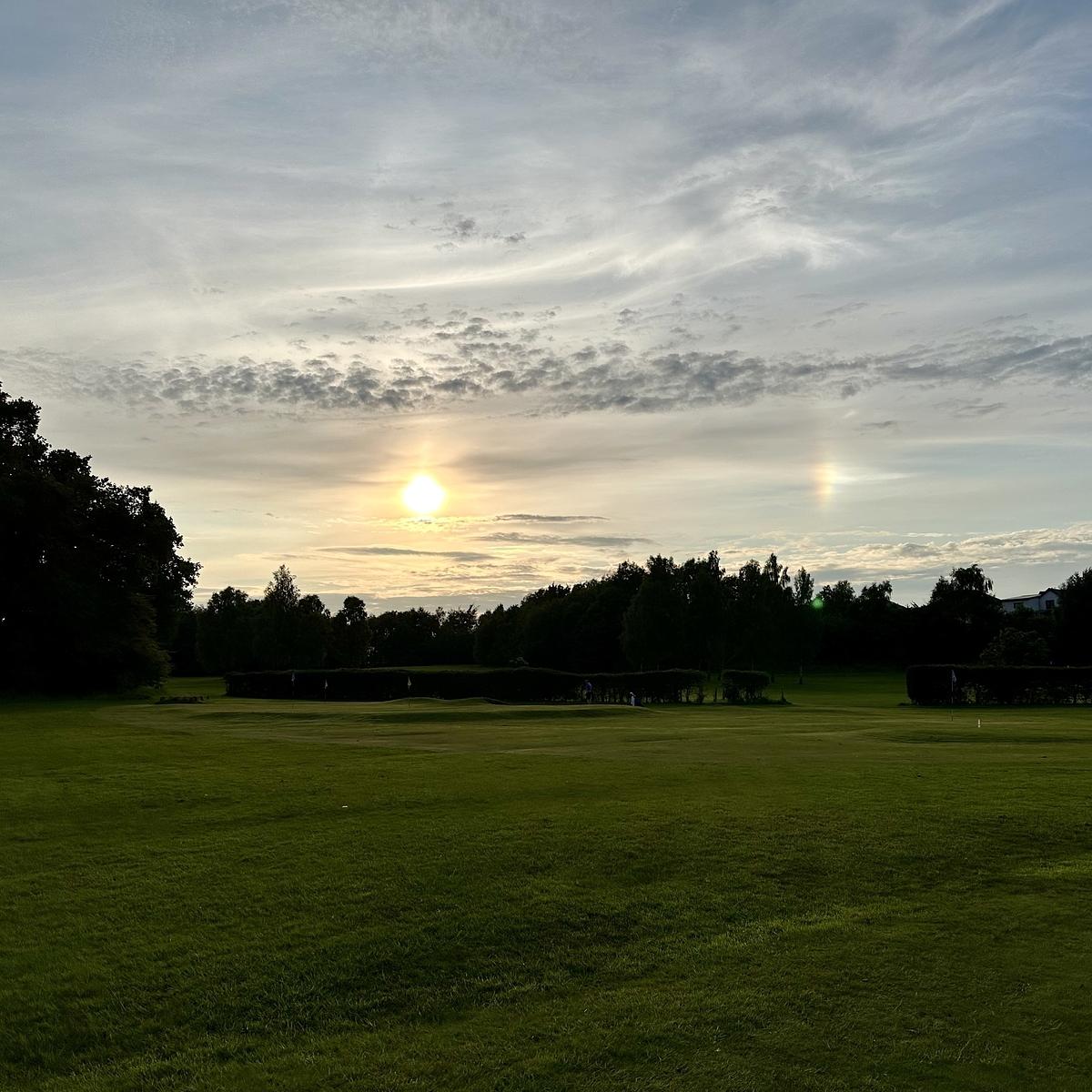 A grassy field stretches out under a partly cloudy sky with a bright sun and a prominent sundog near the horizon.