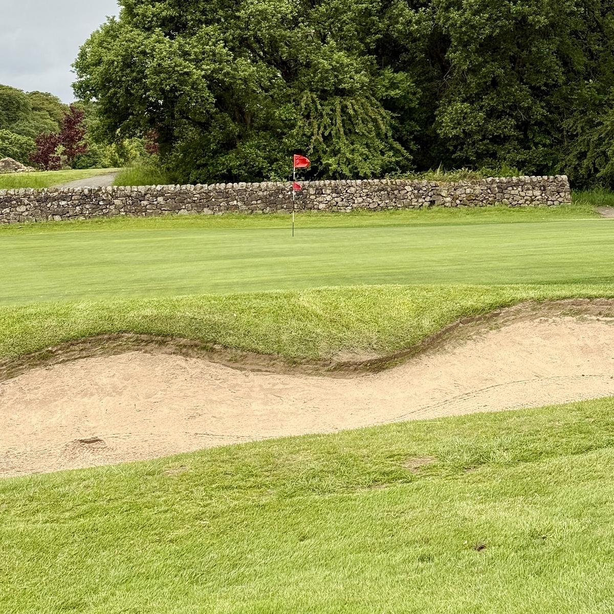 A golf course green with a sand bunker in the foreground and a red flag marking the hole, surrounded by lush greenery and stone walls.