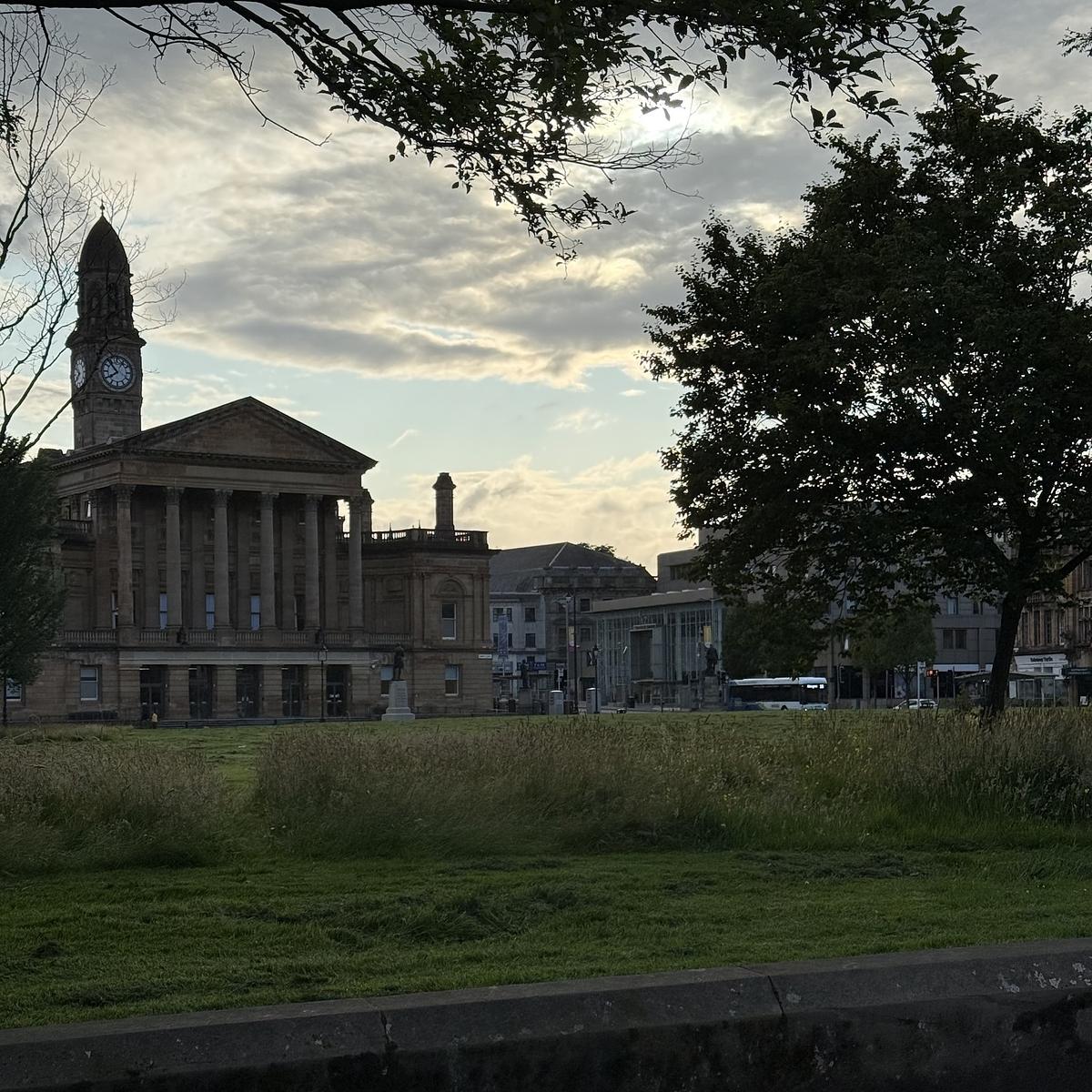 A historic building with columns and a clock tower is surrounded by green lawns and trees under a cloudy sky.