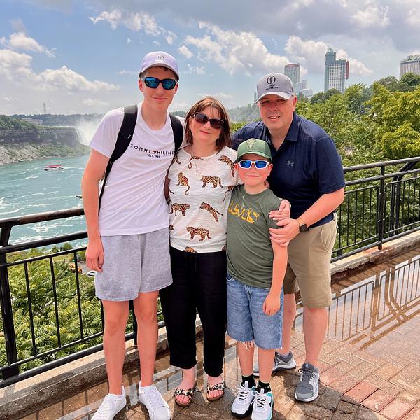 A family of four, consisting of two adults and two children, is posing together on a walkway with a scenic view of a waterfall and city skyline in the background.