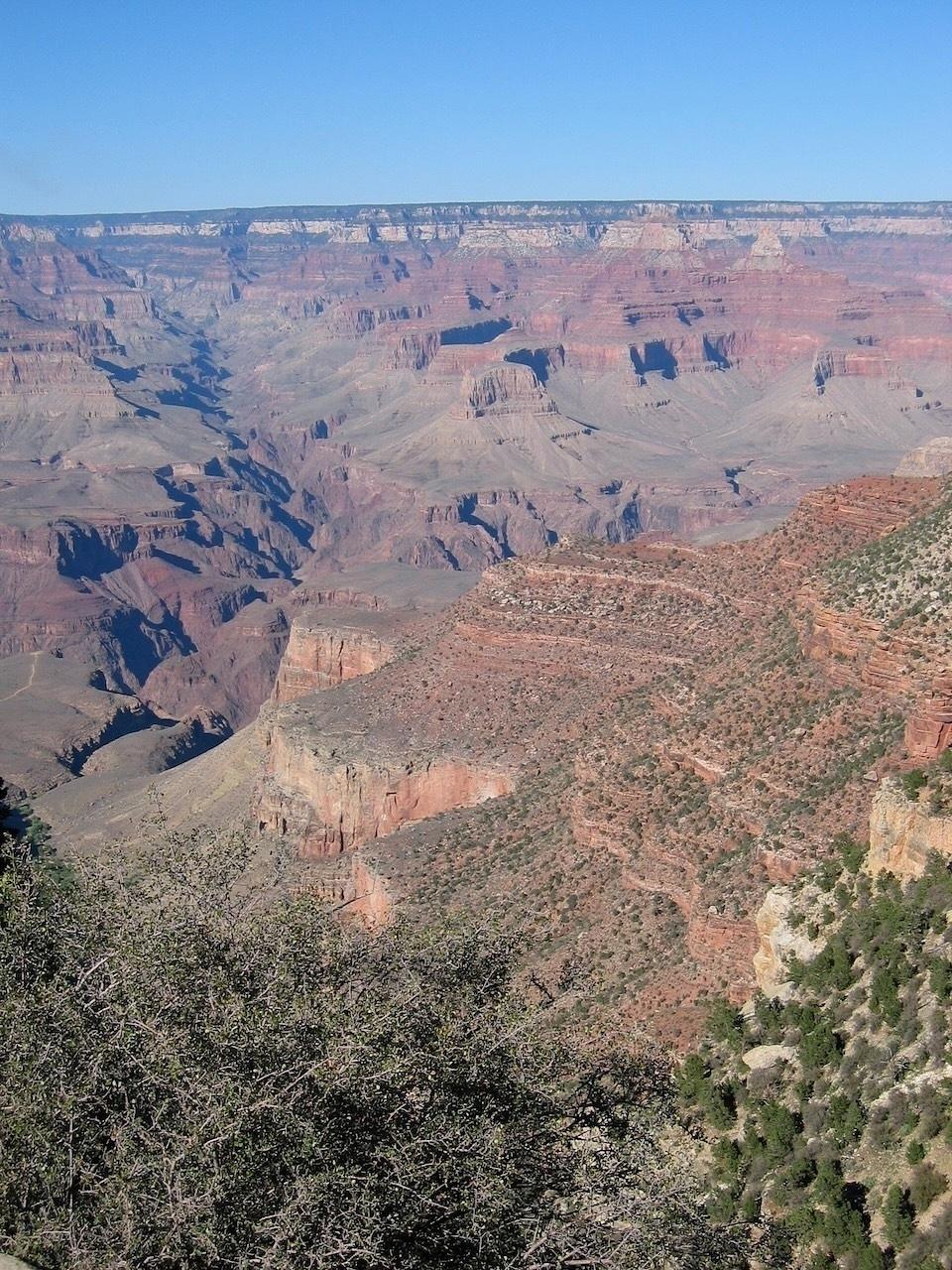 A vast, colorful landscape of the Grand Canyon stretches under a clear blue sky with rugged rock formations and deep valleys.