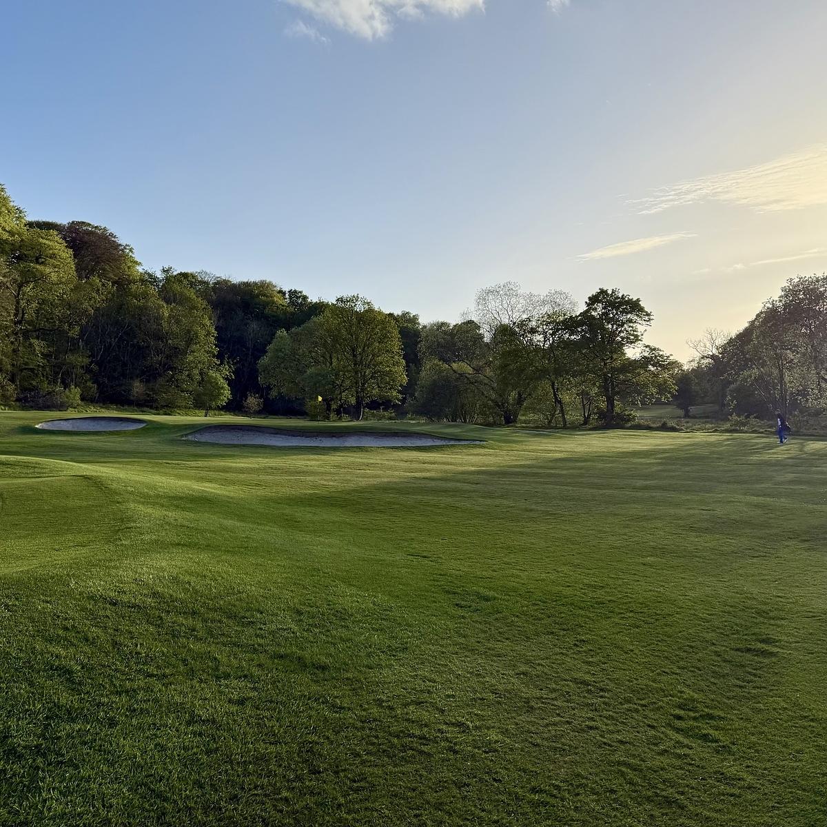 A lush golf course features well-maintained greens, sand bunkers, and is surrounded by trees with a person visible in the distance.