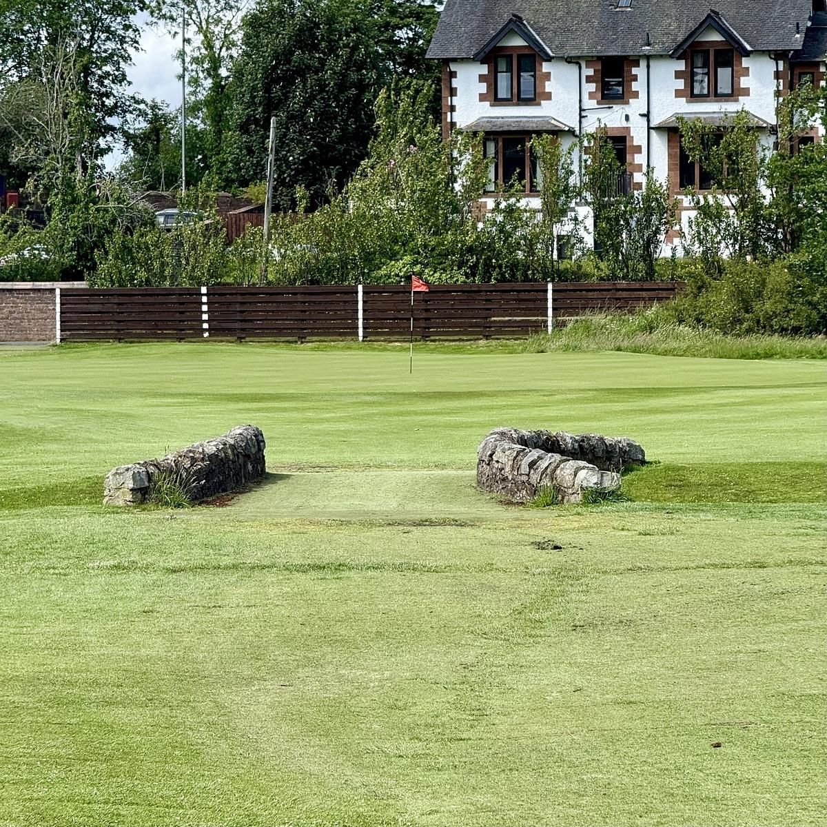 A manicured golf green with a flagstick is bordered by stone markers, trees, and a house in the background.