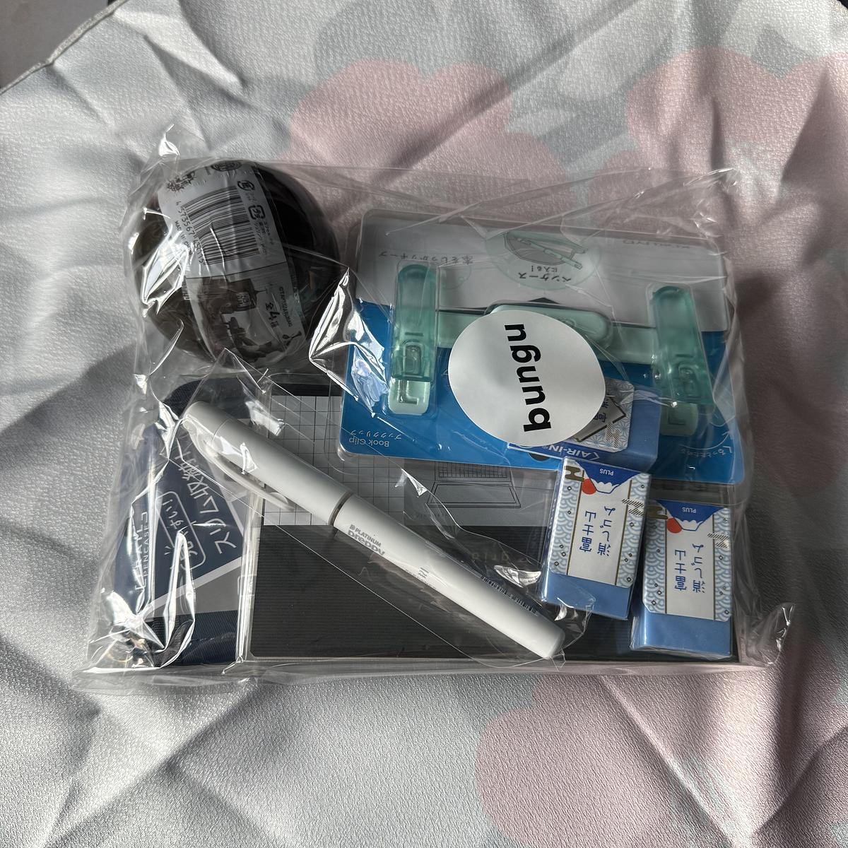 A clear plastic bag of Japanese stationery items labelled "bungu," containing various office supplies including pens, erasers (PLUS brand), a book clip, washi tape, and other writing accessories, displayed on a light fabric surface.