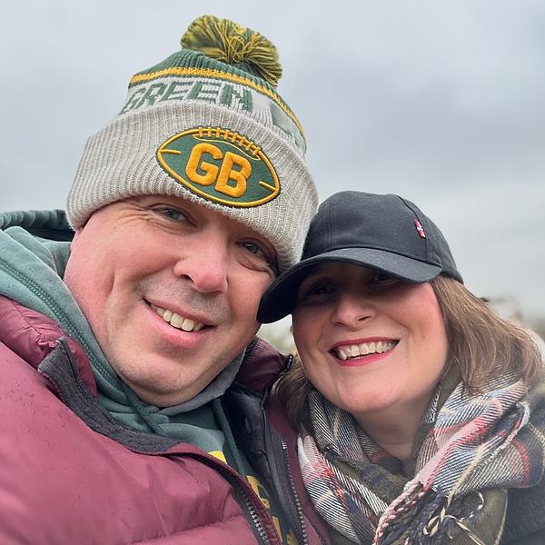 A smiling couple wearing winter clothing, including a Green Bay Packers beanie and a black cap, pose for a selfie outdoors.