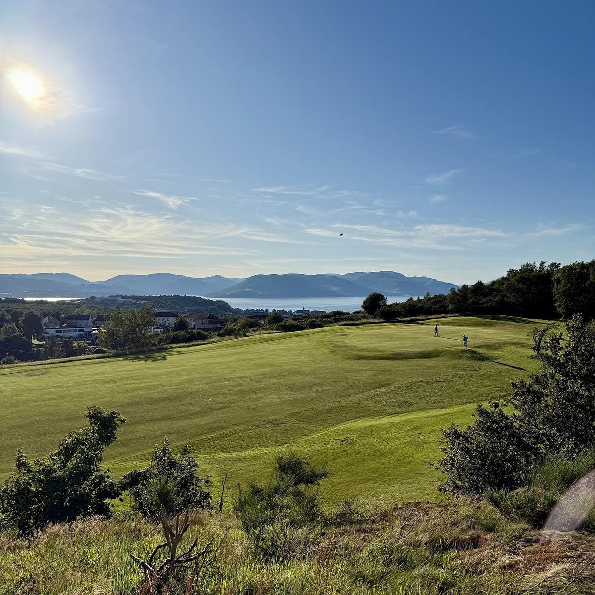 A scenic landscape features a lush green golf course, trees, and a hilly backdrop under a bright, sunny sky.