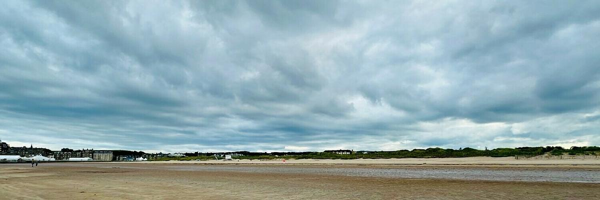 Auto-generated description: A vast, empty beach stretches out under a cloudy sky with a few buildings and greenery in the distance.