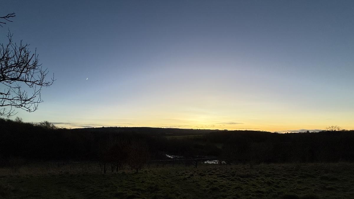 A serene landscape at dusk features a silhouetted horizon with a subtle glow in the sky and a small crescent moon to the left.
