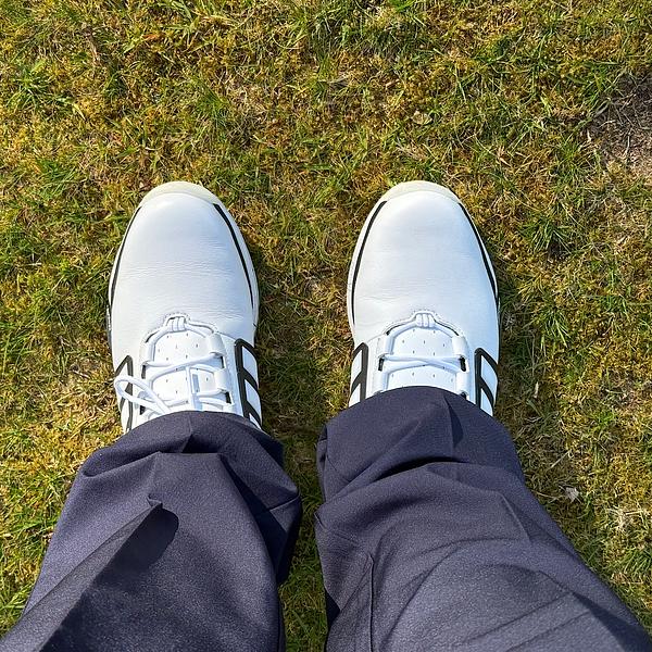 A person looking down at their white and black golf shoes while standing on green grass, wearing dark navy trousers.
