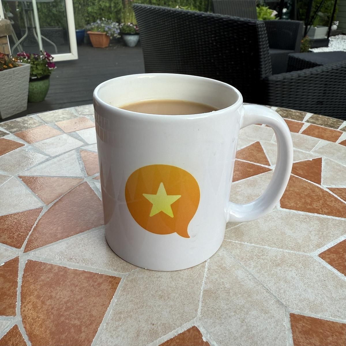 A white mug with a star inside a speech bubble design sits on a mosaic-patterned outdoor table, holding a light-colored beverage.