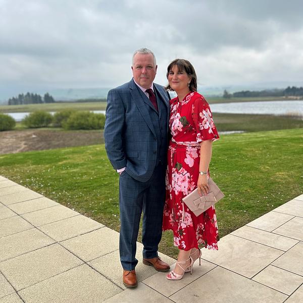 A man in a blue suit and a woman in a red floral dress stand together on a paved area with a scenic background of grass, water, and cloudy skies.
