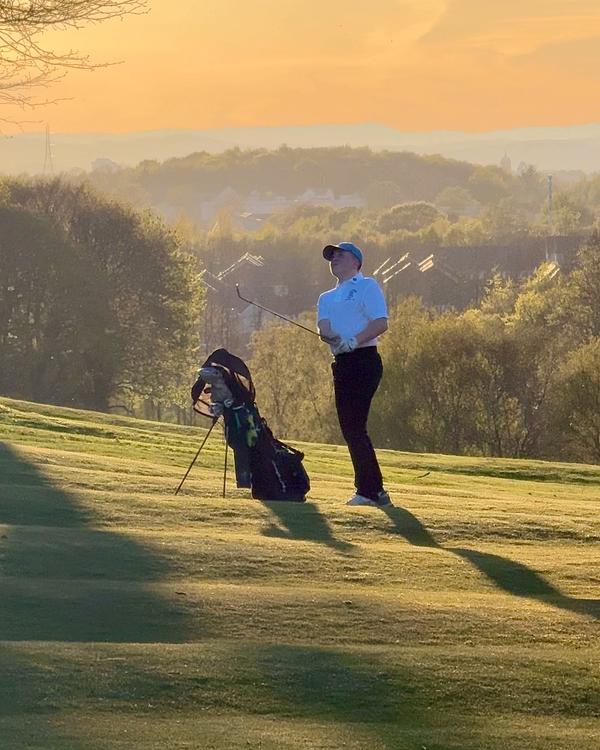A golfer standing on a green hillside at sunset, holding a club and watching their shot, with a golf bag nearby.