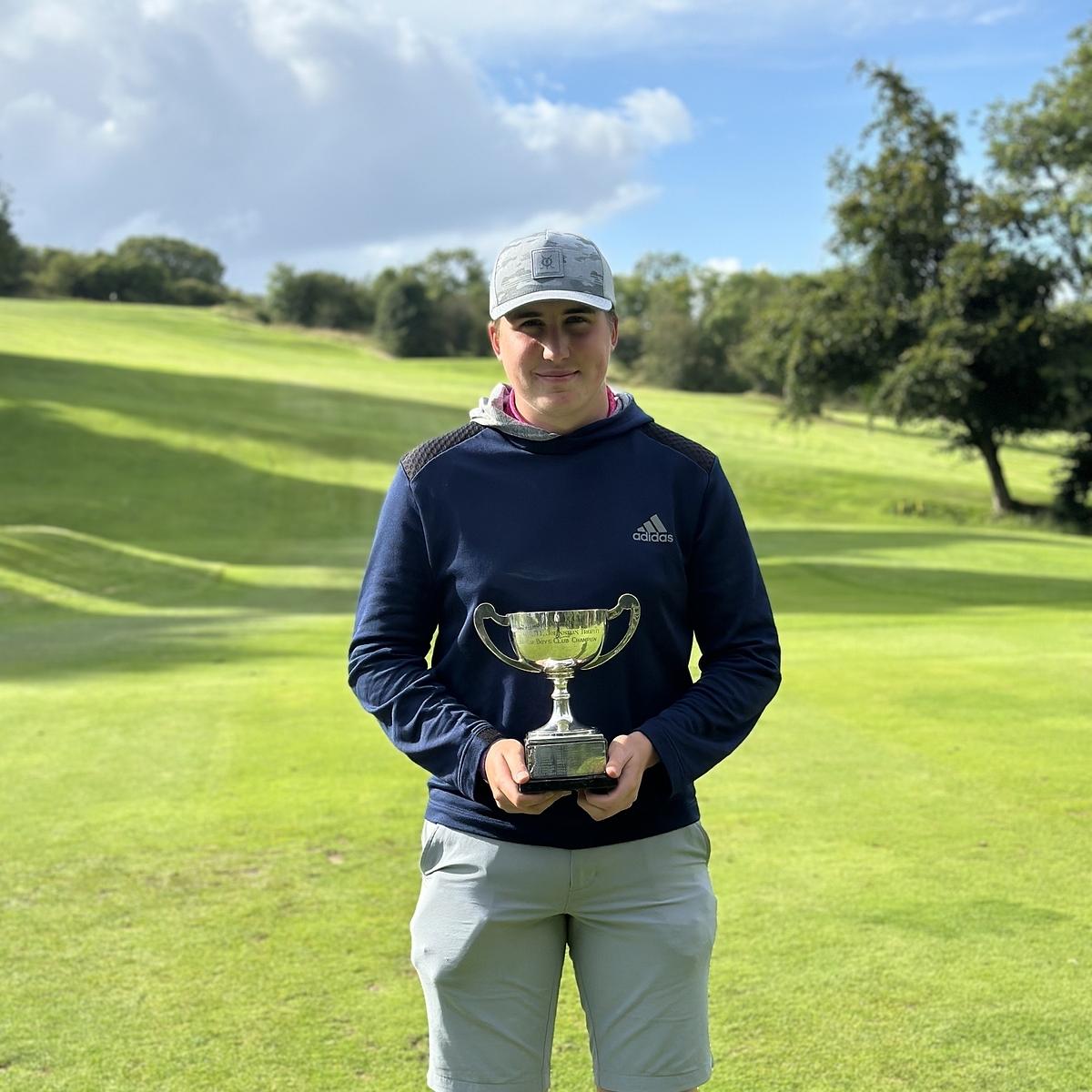 A person is standing on a golf course holding a trophy, with trees and a cloudy sky in the background.
