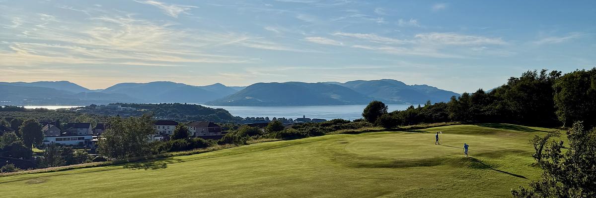 Auto-generated description: Golfers on a lush green course overlooking a coastal town, calm sea, and misty mountain range under a soft evening sky.