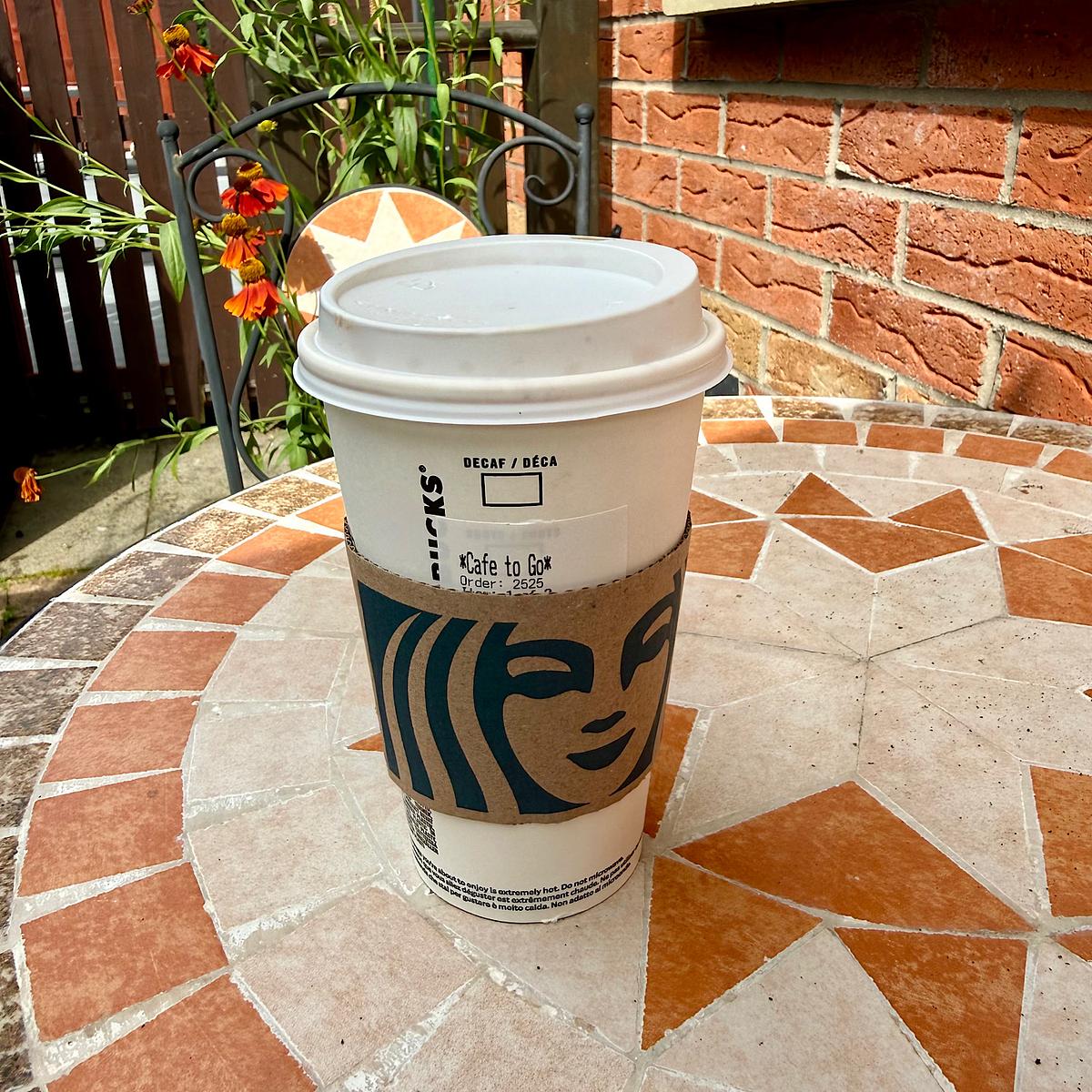 A Starbucks takeout coffee cup on a mosaic patio table, with orange flowers and a brick wall in the background.