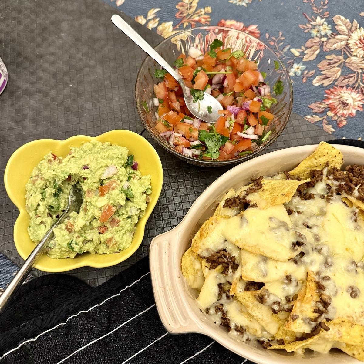 A dish of cheesy nachos with ground beef is accompanied by bowls of guacamole and fresh salsa on a textured placemat.