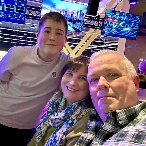 A family of three—a teen boy, woman, and man—posing for a selfie at a bowling alley with purple lighting and scoreboards.