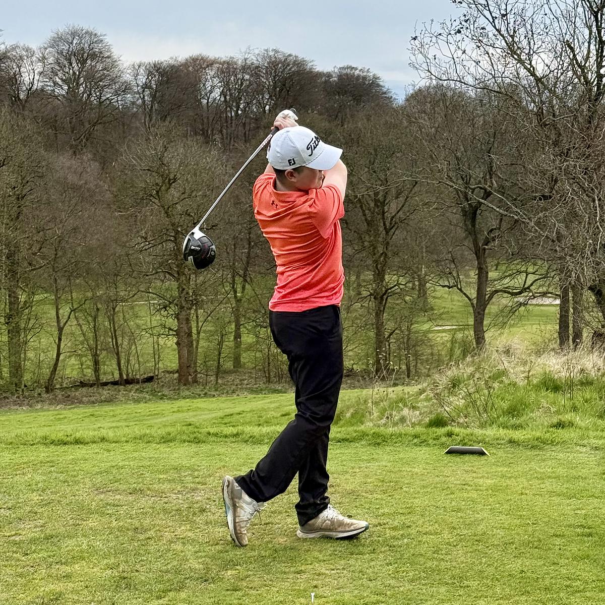 A golfer in an orange shirt and white Titleist cap mid-swing on a tee box, with leafless trees in the background.