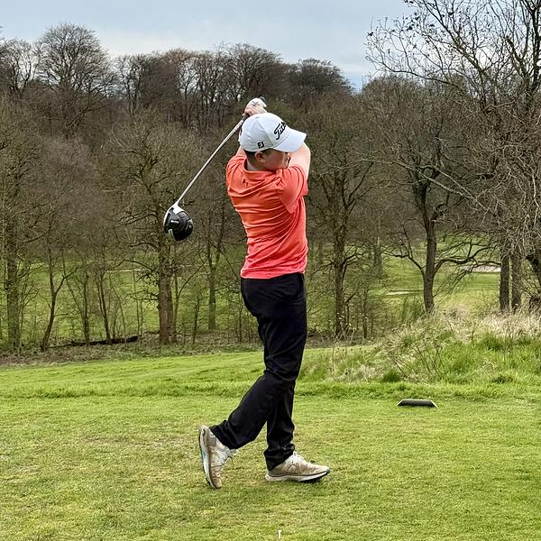 A golfer in an orange shirt and white Titleist cap mid-swing on a tee box, with leafless trees in the background.