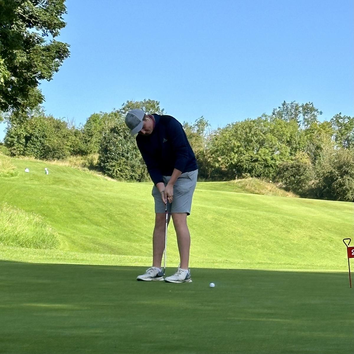 A man in a navy shirt and grey shorts is putting on a golf green surrounded by trees and grassy hills.