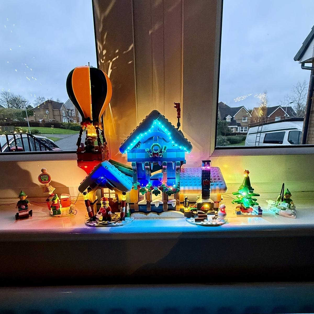 A festive holiday scene with illuminated miniature houses, a hot air balloon, and figurines is displayed on a windowsill.