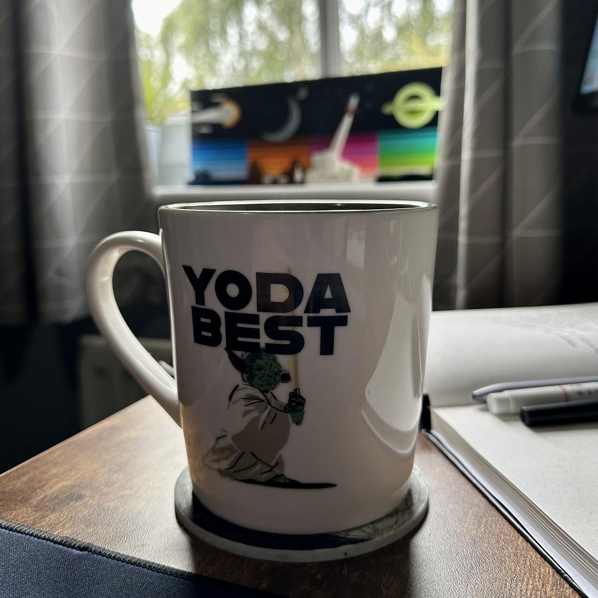 A mug with "YODA BEST" and an illustration of Yoda is placed on a wooden surface near books and a window.