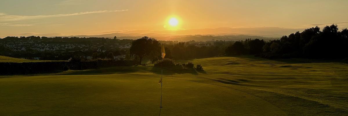 Golf course green with flagstick at sunset, overlooking a residential area with hills in the distant background.