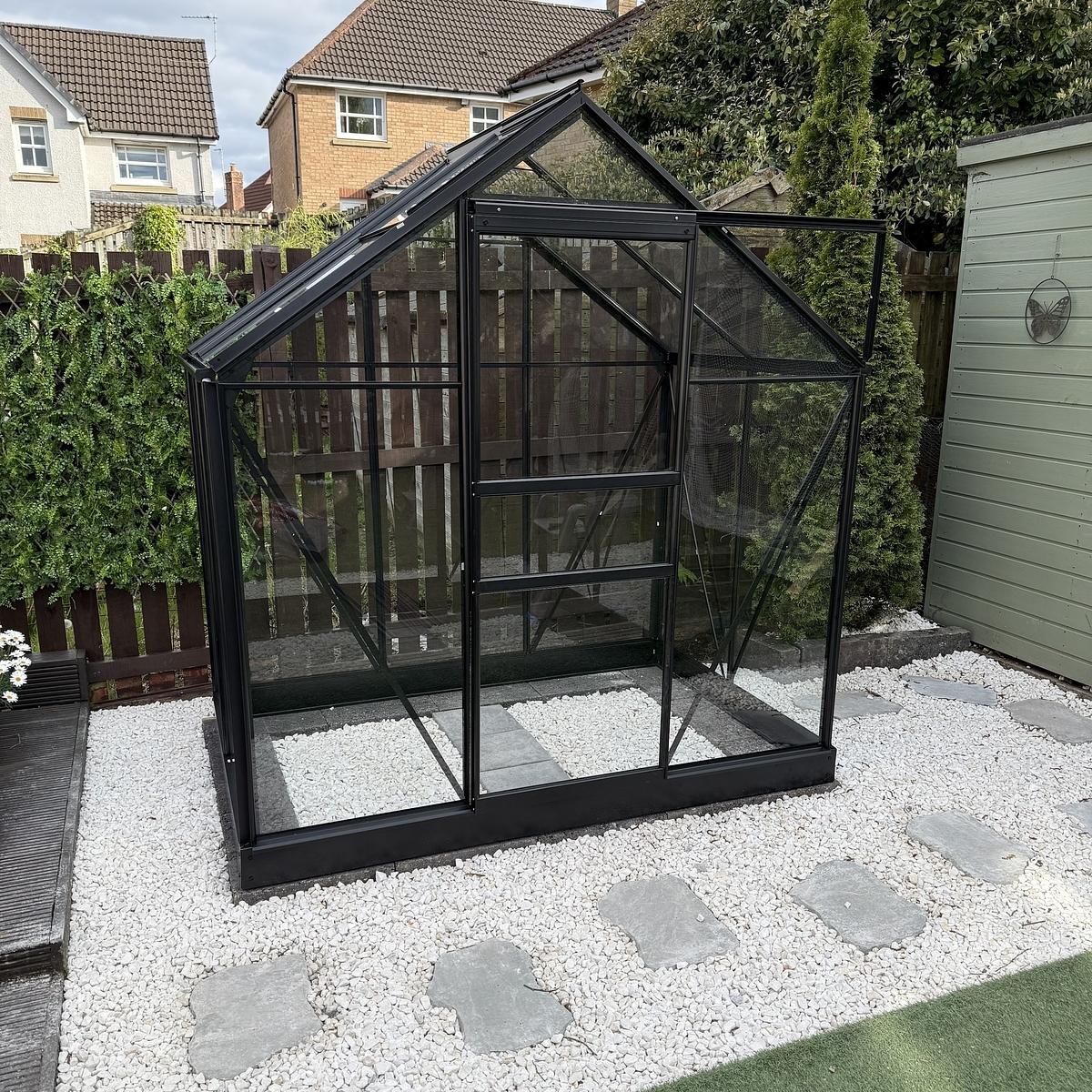 A small glass greenhouse with a black frame is situated on a bed of stones in a backyard garden.