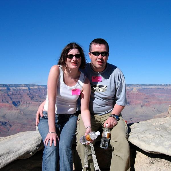 Two people are sitting on a rock ledge with a vast canyon landscape in the background, both wearing casual clothes and sunglasses, each holding a bottle.