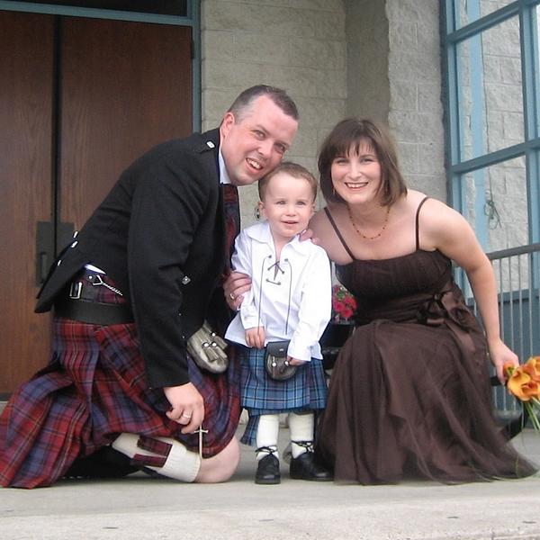 A family poses together, with the father and child wearing traditional Scottish kilts while the mother holds flowers.