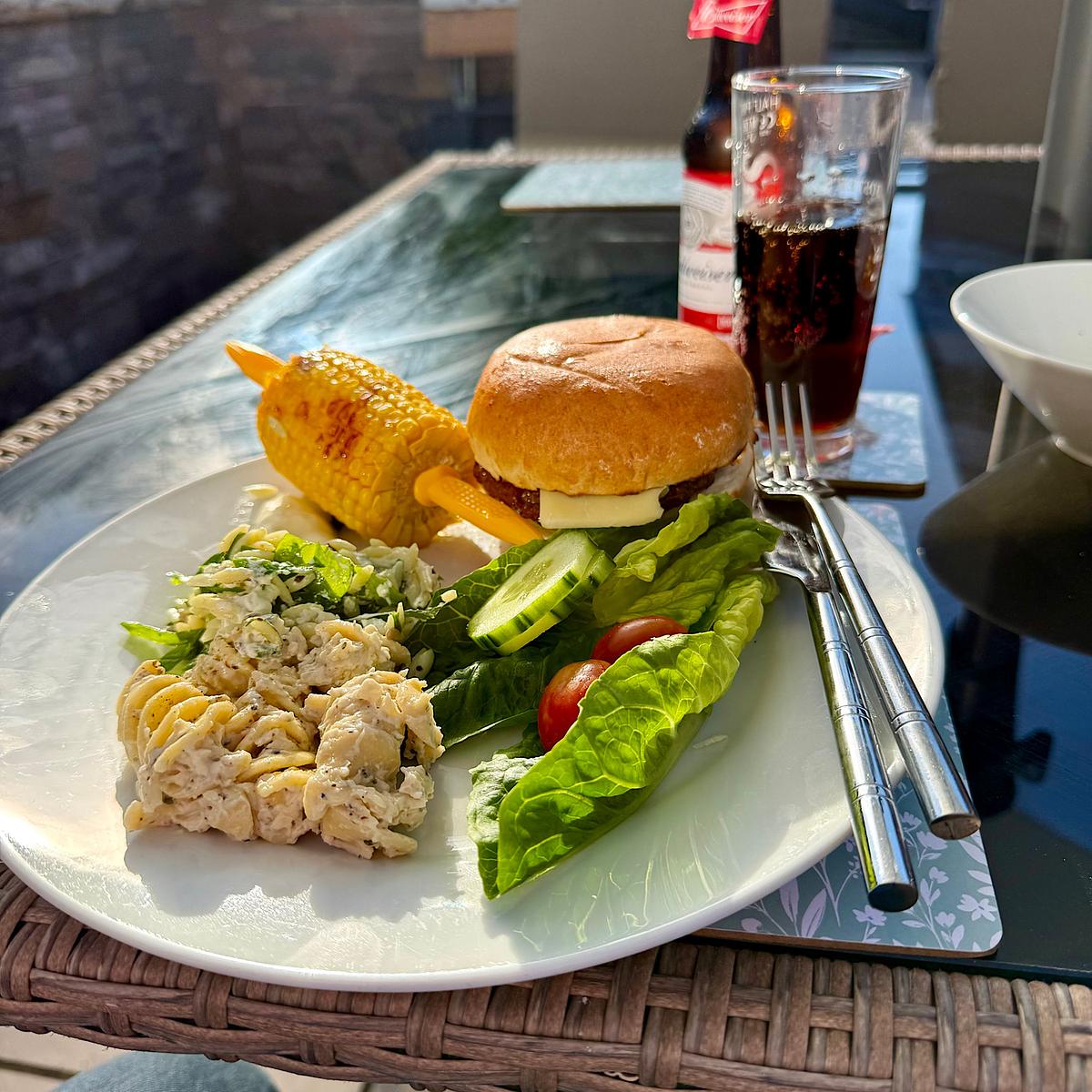 A plate with a burger, grilled corn on the cob, pasta salad, coleslaw, and fresh salad, with a Budweiser beer and cola glass.