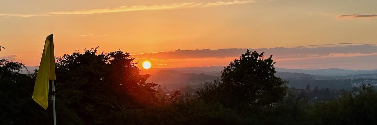 Auto-generated description: Sunset over a hilly landscape with silhouetted trees and a yellow golf flag in the foreground.