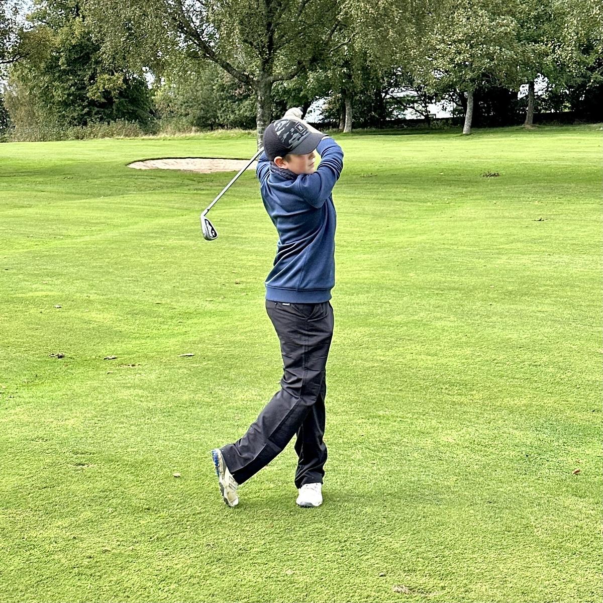 A golfer is completing a swing on a grassy golf course surrounded by trees.