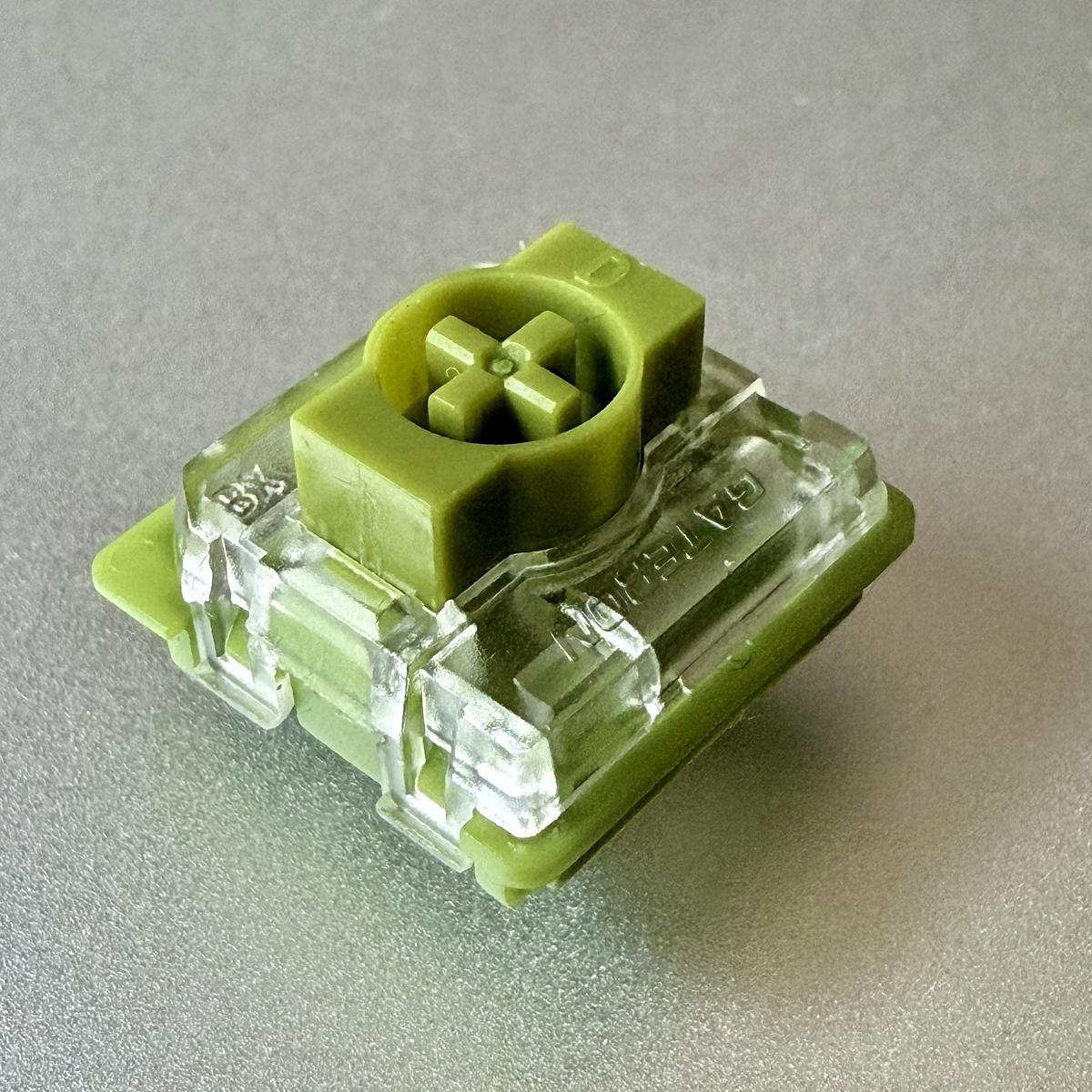 A green mechanical keyboard switch with a transparent housing is placed on a gray surface.