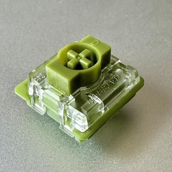 A green mechanical keyboard switch with a transparent housing is placed on a gray surface.