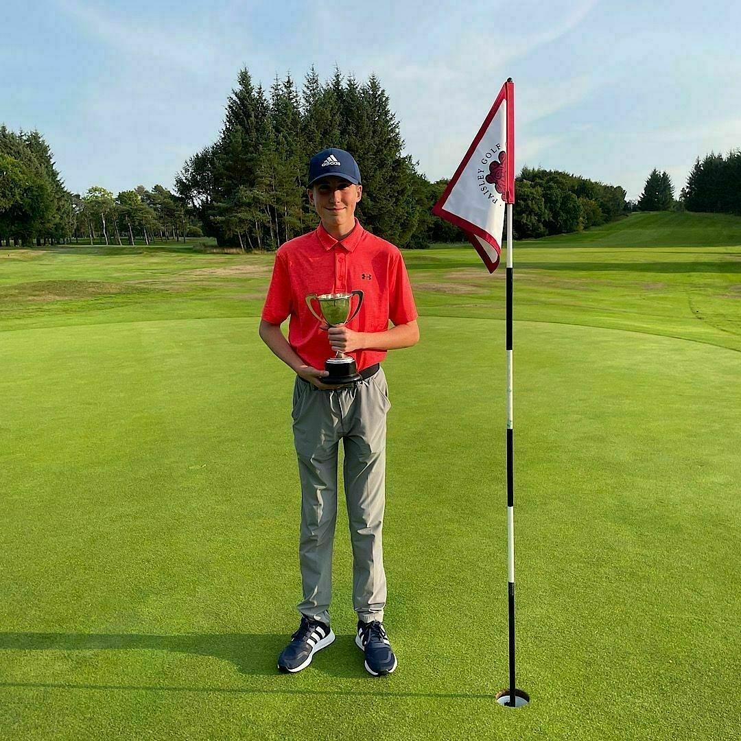 Auto-generated description: A young person in a red shirt and gray pants stands on a golf course holding a trophy next to a flagstick.
