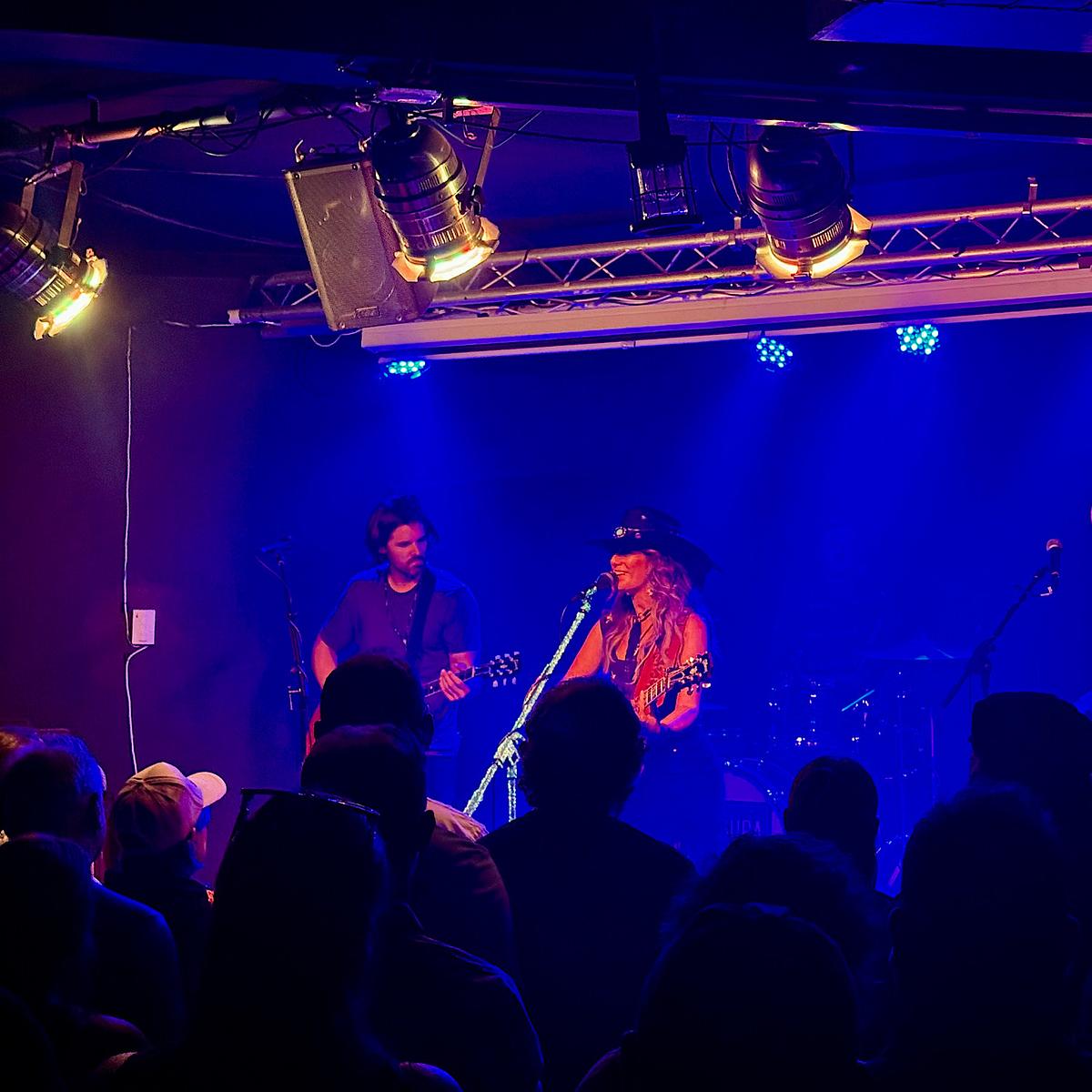 Two musicians performing on stage at an intimate venue bathed in blue light, with a guitarist and a singer wearing a hat, audience silhouettes in foreground.