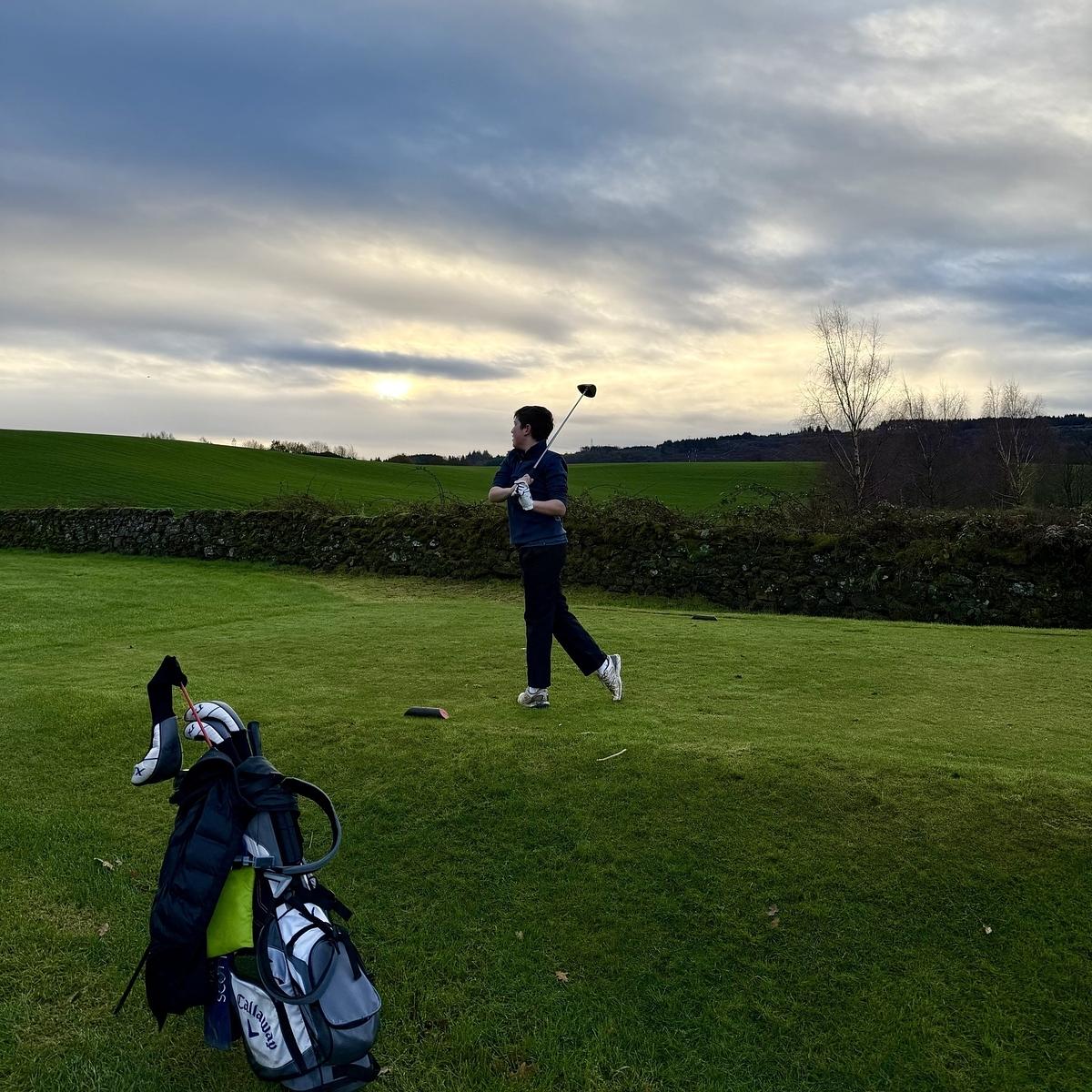A person is golfing on a grassy course under a cloudy sky, with a golf bag in the foreground.