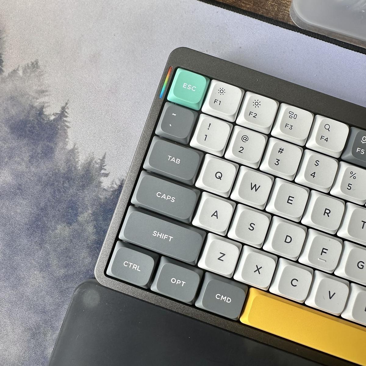 A computer keyboard with a minimalist, custom design features a mix of colorful and monochrome keys, along with a forest-themed mouse pad in the background.