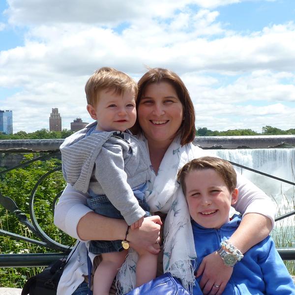 A woman and two children are smiling in front of a waterfall, with buildings visible in the background.