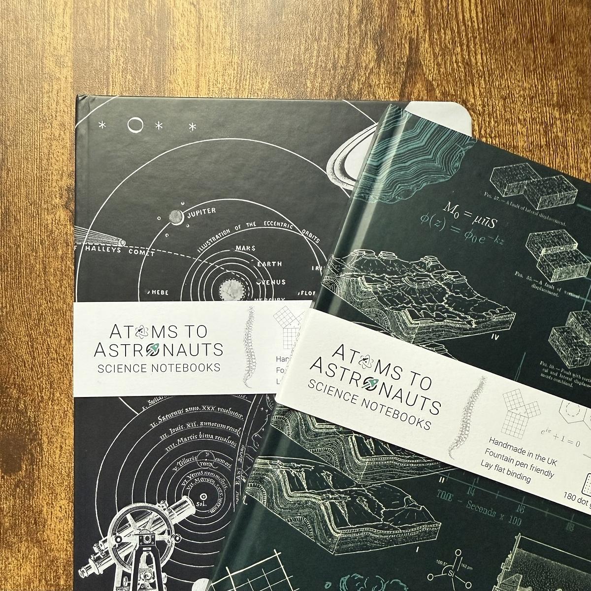 Two science-themed notebooks, one featuring a geological design and the other with planetary orbits, are titled "Atoms to Astronauts Science Notebooks."